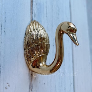 Può includere: Un gancio dorato a forma di cigno con piume dettagliate sul corpo. Il gancio è montato su una superficie di legno bianco.