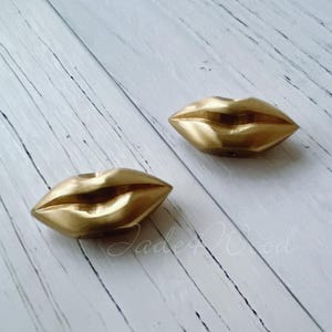 May include: Two gold-coloured metal lips, shaped like a mouth, are shown on a white wooden surface.