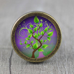 Könnte beinhalten: Ein runder, antiker goldfarbener Schrank-Knopf mit einem violetten und grünen Baum des Lebens-Design.
