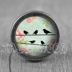 May include: A round, silver knob with a floral design and five black birds on a wire. The floral design features pink roses on a light blue background.