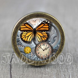 May include: A round, brass-colored knob with a steampunk design featuring a butterfly with orange and black wings, gears, and a clock face.