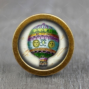 Puede incluir: Un pomo redondo dorado con una imagen vintage de un globo aerostático. El globo es verde, morado y amarillo con un patrón blanco y dorado. El globo tiene una cesta y está volando en el cielo.
