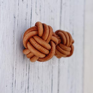 Könnte beinhalten: Zwei braune dekorative Objekte in Form von Lederknoten. Die Knoten sind eng gewebt und haben eine strukturierte Oberfläche. Die Objekte befinden sich vor einem weißen Holzhintergrund. Der Text "Jade4Wood" ist sichtbar.