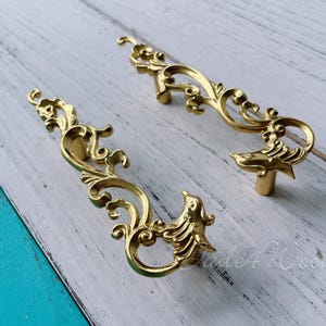 Op de afbeelding: Twee sierlijke, goudkleurige ladegrepen met een decoratief scroll-ontwerp. De grepen hebben een vogelachtige vorm aan de onderkant.