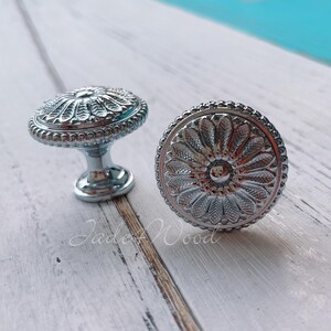 May include: Two silver-toned cabinet knobs with a decorative floral design. The knobs have a raised, ornate pattern and a smooth, shiny finish.