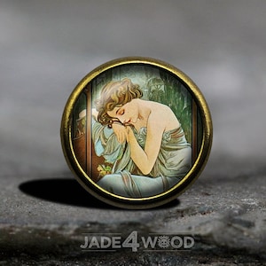 Könnte beinhalten: Ein runder Anhänger aus Messing mit einem Glaseinsatz, der eine Vintage-Illustration einer Frau in einem blau-weißen Kleid zeigt, die schläft und ihren Kopf auf ihrer Hand ruht. Der Hintergrund ist ein grünes und braunes Blumenmuster.