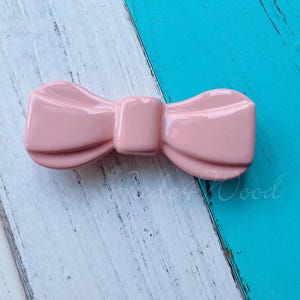 以下が含まれることがあります： ピンクのセラミック製の蝶ネクタイ型のヘアクリップ。クリップは白と青の背景の上にあります。