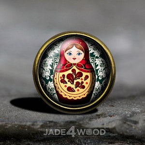 Puede incluir: Un pin redondo de metal dorado con un cabujón de vidrio que presenta una ilustración colorida de una muñeca rusa tradicional. La muñeca es roja, amarilla y verde con una cara blanca y ojos negros. El fondo es un patrón floral verde y blanco.