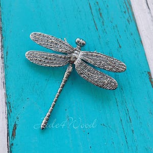 May include: A silver-colored dragonfly brooch with detailed wing patterns rests on a distressed turquoise wooden surface. The dragonfly's body and wings are intricately designed, showcasing a textured finish. The brooch is a decorative accessory.