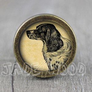 May include: A round, brass-colored knob with a vintage-style illustration of a dog's head in black and white on a cream background.