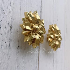 May include: Two gold-toned metal flower-shaped cabinet knobs. The knobs have a detailed, intricate design with a raised center.