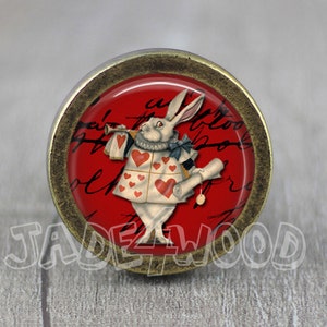 Puede incluir: Un pomo de armario redondo de color latón con una imagen roja y blanca del Conejo Blanco de Alicia en el País de las Maravillas. El conejo lleva un chaleco con corazones y sostiene una trompeta. El fondo es rojo con escritura negra.