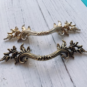 May include: Two ornate, antique-style drawer pulls. One is a bright gold color, the other is a darker bronze. Both have a curved handle with floral and leaf detailing on the ends.