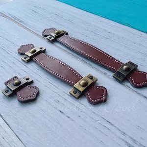 Może przedstawiać: Zestaw trzech brązowych skórzanych uchwytów do szafek z mosiężnym osprzętem. Uchwyty mają szyty wzór i są w różnych rozmiarach. Okucia są nitowane do skóry. Tło to jasnoniebieska i biała drewniana powierzchnia.