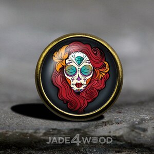 May include: A round, gold-toned metal pin featuring a colourful image of a sugar skull with red hair and flowers. The skull has a diamond-shaped jewel on its forehead.