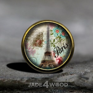 May include: A round, brass-coloured, decorative knob with a glass insert featuring a vintage image of the Eiffel Tower in Paris, France. The image includes flowers, a blue sky, and the word "Paris".