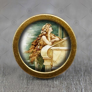 Könnte beinhalten: Ein runder, goldfarbener Metallknopf mit einem kreisförmigen Bild einer Meerjungfrau mit langen, fließenden Haaren, die auf ein grünes und blaues Meer blickt. Die Meerjungfrau sitzt auf einer weißen Steinleiste.