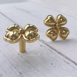 May include: Two gold-toned metal flower-shaped cabinet knobs. The knobs have five petals each and a small center piece. The knobs are on a white wooden surface.