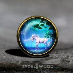 May include: A gold-toned metal button with a round, glass cabochon featuring a white unicorn with a blue horn and a blue and green forest background.
