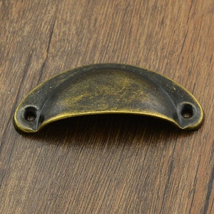 May include: Antique brass cabinet pull with a rounded, slightly curved shape. The pull has two small holes for mounting.