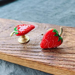 May include: Two gold-coloured strawberry-shaped drawer pulls with red and green enamel. The strawberries are on a wooden surface.