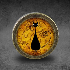 Puede incluir: Un pomo redondo de color latón con una silueta negra de un gato sobre un fondo amarillo con diseños florales.