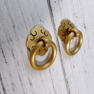 Puede incluir: Dos tiradores dorados para armarios con una placa trasera decorativa en forma de nube y un tirador de anillo. Los tiradores están montados sobre una superficie de madera pintada de blanco, creando un contraste con los cálidos tonos dorados.