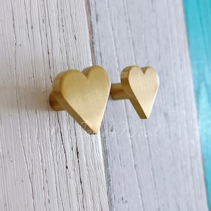 Puede incluir: Dos pomos de armario en forma de corazón de color dorado, uno más grande que el otro, montados en una superficie de madera blanca.