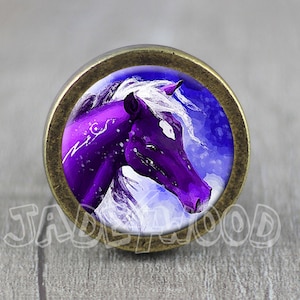 Op de afbeelding: Een paars paardenhoofd met een witte manen en staart, geschilderd op een ronde glazen cabochon, ingelijst in een bronzenkleurig metalen frame.