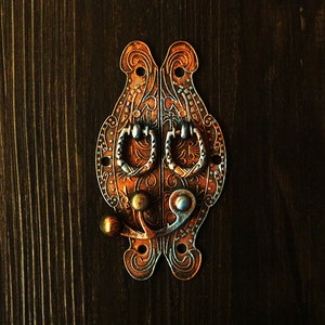 Puede incluir: Pestillo de metal adornado con dos manijas decorativas y un diseño floral. El pestillo es de color cobre oscuro con detalles plateados.