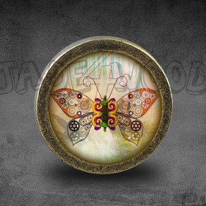 Op de afbeelding: Een ronde, messingkleurige knop met een decoratief, steampunk-stijl vlinderontwerp. De vlinder is gemaakt van tandwielen en andere mechanische elementen en heeft een kleurpalet van paars, groen en oranje.