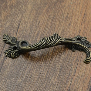 Puede incluir: Una manilla de tiro de metal de estilo antiguo, de color bronce, con un diseño de hojas. La manilla tiene un patrón de espiral decorativo y dos agujeros para su montaje.