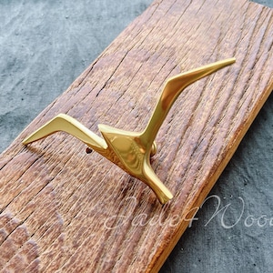 May include: A gold-coloured metal bird-shaped pin on a weathered wooden plank. The pin has a sleek, modern design with outstretched wings. The wood has a natural, aged appearance, with visible grain and texture.