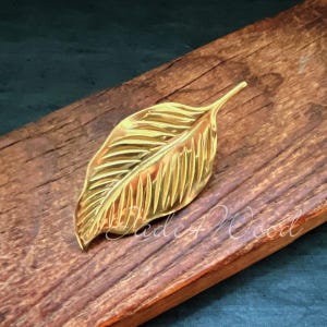 Könnte beinhalten: Ein goldfarbener, blattförmiger Türgriff mit strukturierter Oberfläche. Der Griff befindet sich auf einer Holzoberfläche.