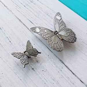 May include: Two metal butterfly drawer pulls in a distressed bronze finish. The larger butterfly has a wingspan of approximately 7.5 cm. The smaller butterfly has a wingspan of approximately 5 cm.