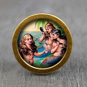 May include: A gold-toned round pin with a colorful image of three mermaids with long flowing hair. The mermaids are wearing seashells and are holding a bottle and a mirror. The background is a blue ocean with a palm tree.