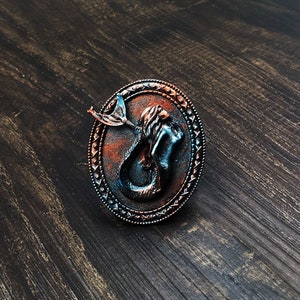 Può includere: Una statuetta di sirena in rame e blu con un design dettagliato, su uno sfondo di legno scuro.