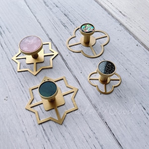 May include: Four gold-toned cabinet knobs with geometric designs. Each knob features a different coloured stone insert: pink, green, black, and iridescent.