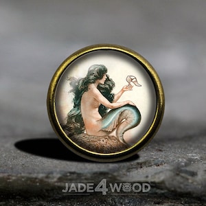 Op de afbeelding: Een ronde gouden knop met een witte achtergrond waarop een zeemeermin met lang groen haar te zien is die een witte schoen vasthoudt. De zeemeermin zit op een rots met een blauwe staart.