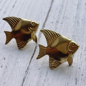 May include: Two gold-coloured metal fish figurines. The fish are stylised and have a detailed design. They are both facing the right side of the image.