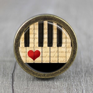 May include: A round, antique gold knob with a glass insert featuring a black and white piano keyboard with a red heart on the keys. The background is a vintage-style paper with text that reads "epite ony, th emboet iesa n nialitie abre and texure. The word der From an art form who medium and and silence An elements ar bite whi"