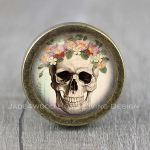May include: A round, antique-style cabinet knob featuring a detailed illustration of a human skull adorned with a floral crown of pink and white flowers. The knob has a bronze-colored metal frame and the text "JADE4WOOD HOME LIVING DESIGN".