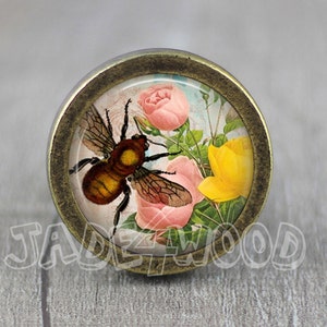 Può includere: Un pomello rotondo color ottone con un inserto in vetro che presenta un'illustrazione vintage di un'ape e fiori rosa e gialli.