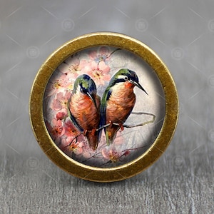 Peut inclure: Un bouton rond doré avec une image colorée de deux oiseaux perchés sur une branche avec des fleurs roses. Les oiseaux ont des plumes bleues, vertes et brunes.