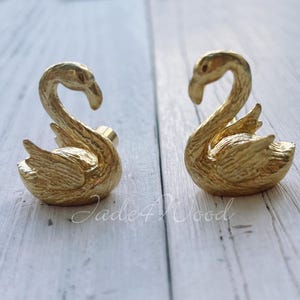 Puede incluir: Dos figuras de cisnes dorados. Los cisnes se enfrentan, con cuellos curvos y texturas detalladas de plumas en sus alas y cuerpos. Estas figuras decorativas son probablemente para la decoración de interiores.