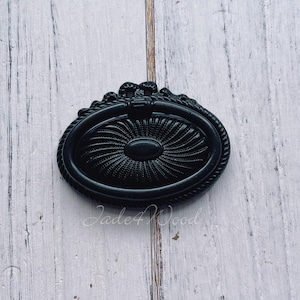 May include: Black oval cabinet pull with a decorative design. The pull has a ring handle and a raised centre.