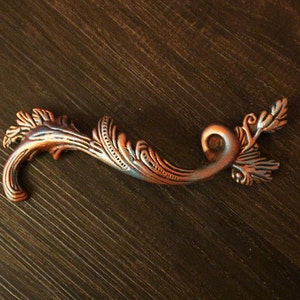Puede incluir: Un tirador de armario decorativo, adornado, de color cobre con un diseño floral.