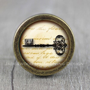 May include: A round, brass-coloured knob with a vintage key design. The key is black and has an ornate design. The knob has a white background with a handwritten message that reads "mes the flas pisrin, and gin advan lage Et And if it does n bau situ".