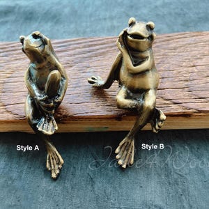 May include: Two brass frog figurines, one sitting with its hands clasped under its chin and the other sitting with its legs crossed. The frog on the left is labeled "Style A" and the frog on the right is labeled "Style B".
