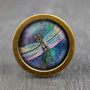 Puede incluir: Un pomo redondo dorado con un inserto de vidrio que presenta una libélula colorida sobre un fondo de acuarela azul y morado con notas musicales y el texto "p.u. dim."